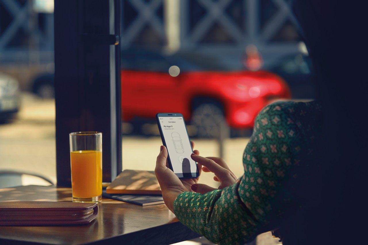 Dáma sedí v kaviarni a používa aplikáciu MyToyota na svojom telefóne, v pozadí je zaparkovaná jej Toyota Yaris Cross.